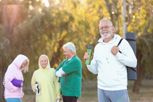 Sporty senior man with fitness mat and water bottle against group in park