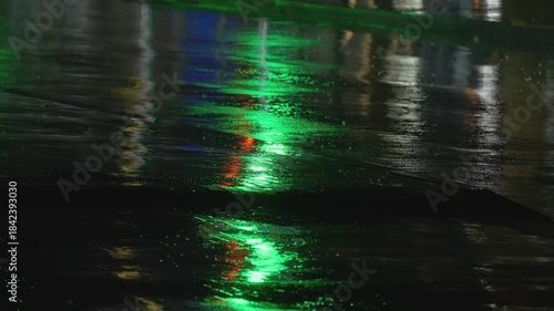 Heavy rain hits the wet asphalt as vivid red and amber reflections from traffic lights and passing cars ripple across the surface. A close-up urban night scene with intense color and moody atmosphere.