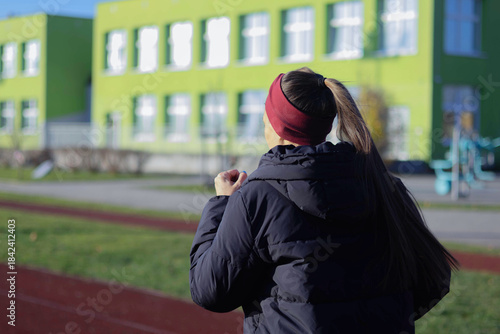 A woman jogs on a sports field by a colorful school building in bright sunlight