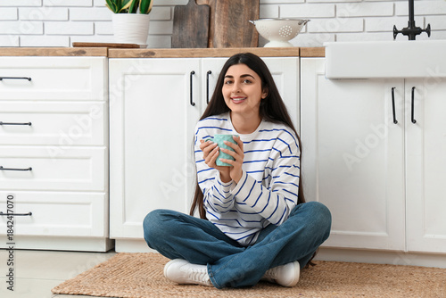 Young woman sitting on kitchen floor with mug, smiling at home in cozy modern...