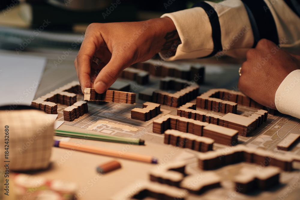 Fototapeta premium Young Black man arranging architectural model pieces on table, hand placing miniature building on urban planning layout, visible tattoo on wrist, pencils and eraser nearby