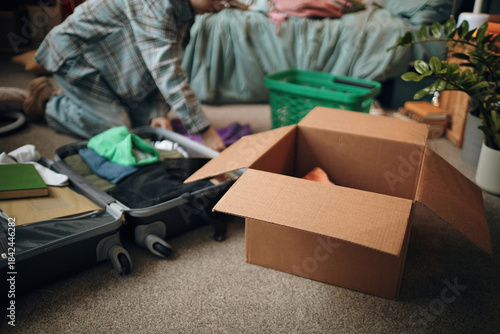 Wallpaper Mural Young woman kneeling on floor packing suitcase with clothing and personal items, open cardboard box and laundry basket nearby, preparing for travel or moving Torontodigital.ca