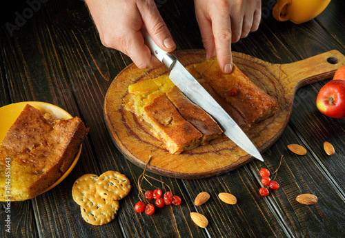 A person cuts a homemade dessert on a wooden board. Nearby are slices of apples, nuts, and crackers. The scene captures a warm, inviting kitchen atmosphere