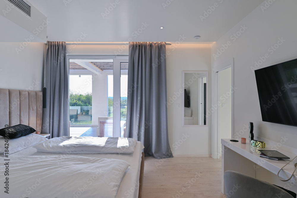 Fototapeta premium Bright, modern bedroom featuring a comfortable bed, sleek desk, and a large sliding glass door opening to a private balcony with stunning green landscape views, perfect for relaxation and serene