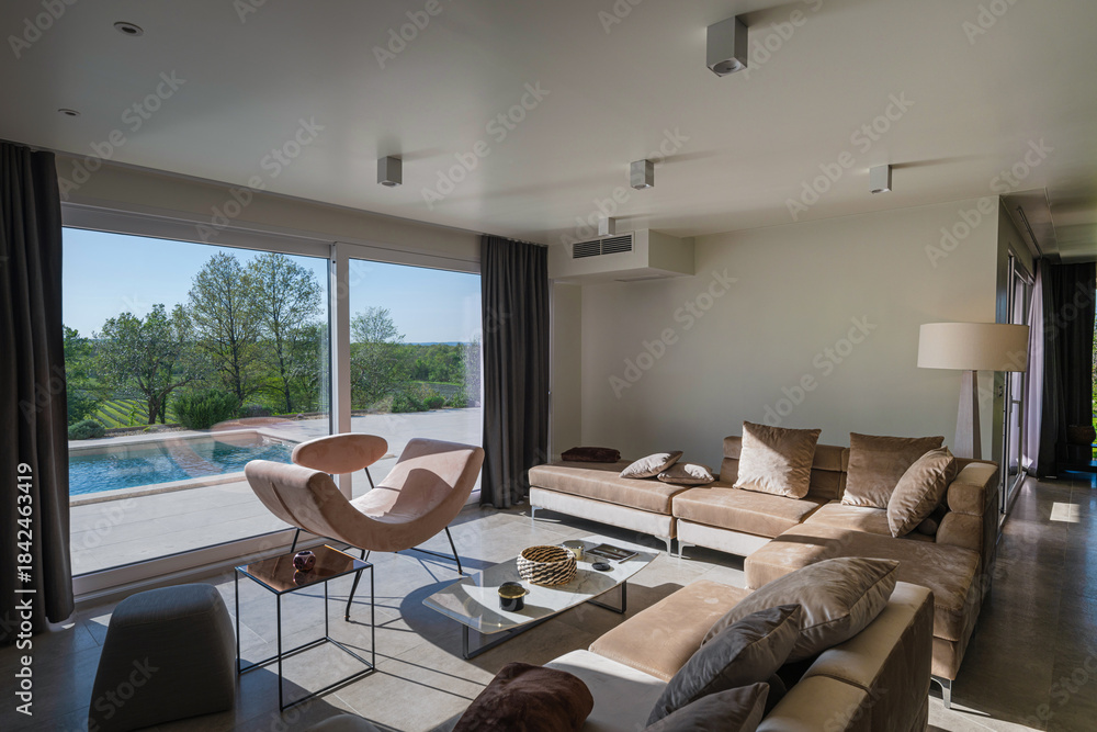 Obraz premium Modern luxury living room interior at sunset. Open-plan space featuring beige sofas, marble dining table, large glass windows with sun flare, and view of a patio. Stylish contemporary home design