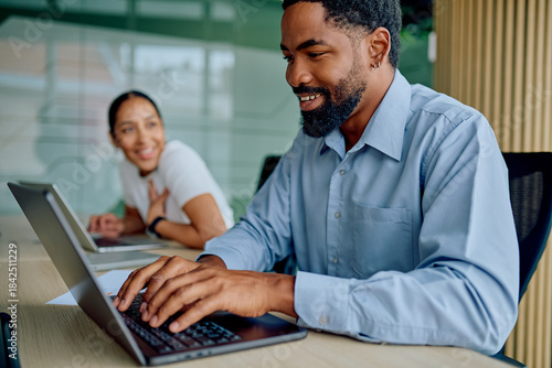 Fotografie Young businessman typing on laptop, smiling in an office while a female colleagu