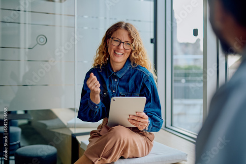 Smiling businesswoman sitting in a modern office, engaged in a conversation while holding a digital tablet