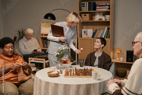 Group of seniors including Black woman knitting, Caucasian man using smartphone, Caucasian woman playing chess with senior man, young adult female nurse offering medication in nursing home