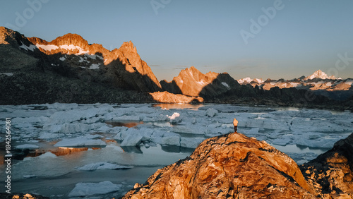View of the lone figure standing atop a sun-kissed rock, overlooking the glacial lake scattered with ice floes and rugged mountains, Ticino, Bedretto, Ticino, Switzerland.