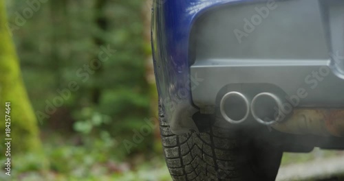 Generic car twin exhaust pipe tips emitting gray smoke or condensation while revving the engine. Low angle, close up shot, blurry nature background, dynamic track and pan real time video, no people