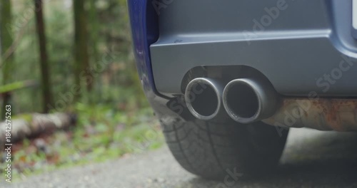 Generic car twin exhaust pipe tips emitting gray smoke or condensation at low engine revs. Low angle, close up shot, blurry nature background, dynamic track and pan real time video, no people