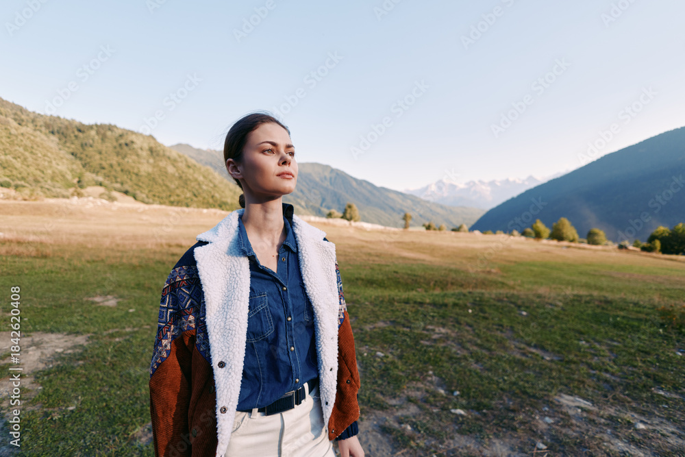 Fototapeta premium Woman portrait in mountains and nature on a valley field, wearing a jacket and denim shirt for travel and outdoors lifestyle, thoughtful pose with scenic horizon and sunlight.