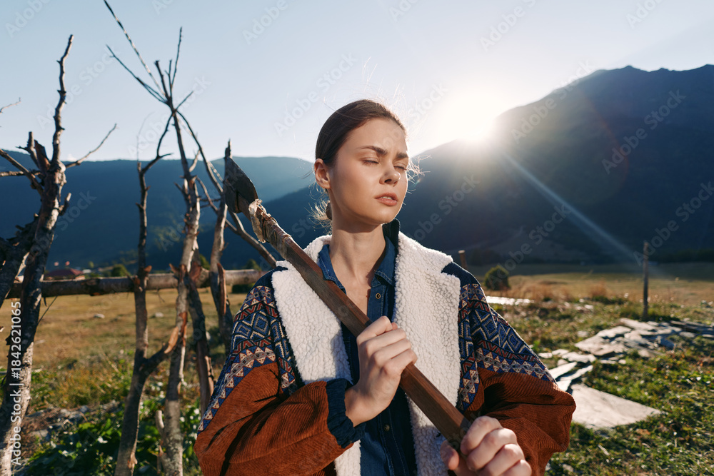 Fototapeta premium woman outdoors mountains sunrise rural stick jacket nature A young woman in a warm jacket holds a wooden stick in a sunlit field, eyes closed and serene against distant mountain peaks.