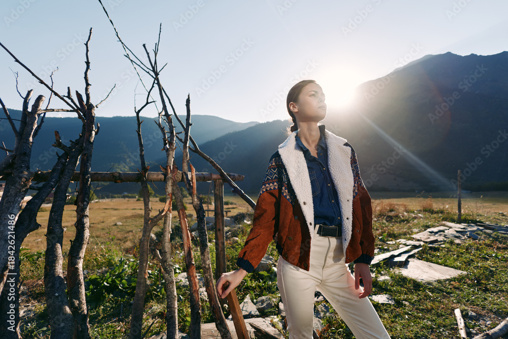 Fototapeta premium Woman in patterned jacket standing in a field with mountains and warm sunlight, posing near rustic wooden sticks and rocks, fashion portrait and lifestyle in natural outdoor landscape.