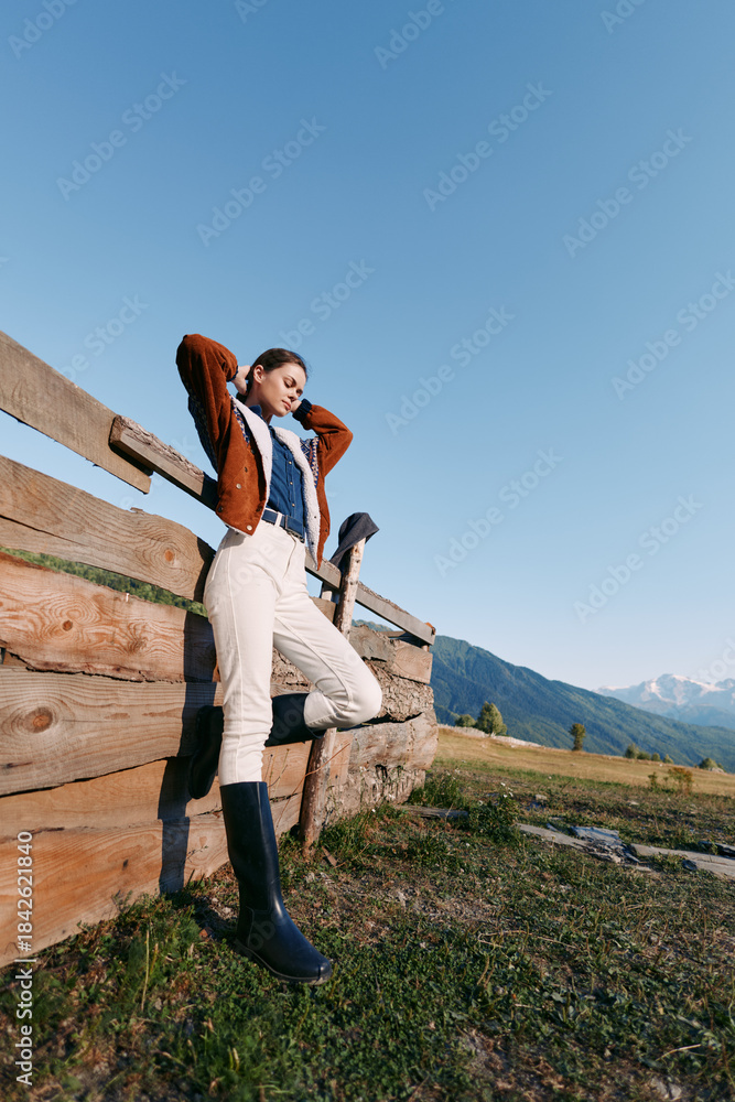 Fototapeta premium Woman by wooden fence in mountains and countryside, wearing boots and jacket in a meadow, portrait pose enjoying sunlight and serene outdoor landscape with blue sky.