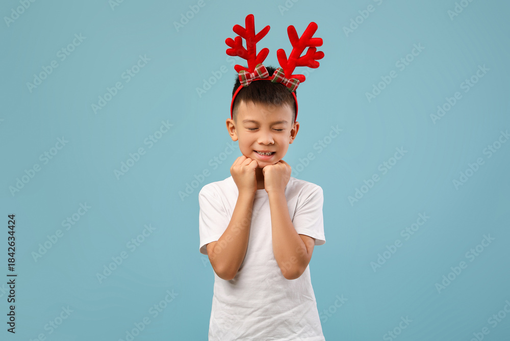 Naklejka premium A joyful young boy stands against a blue background, wearing vibrant reindeer antlers. He closes his eyes and smiles, showcasing pure excitement for the holiday season.