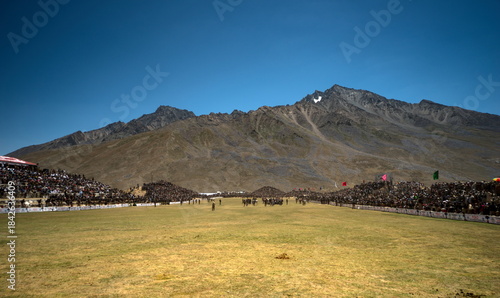 Shandur, Pakistan - 31 July 2017: View of a vast, sun-drenched polo field teeming with spectators against the backdrop of rugged mountains and a clear, azure sky.