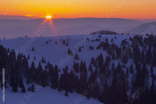 View of the sun's fiery kiss melts the horizon, casting golden rays over the snow-laden peaks and dark forests, a serene winter wonderland, Low Tatras, Banskobystricky kraj, Slovakia.