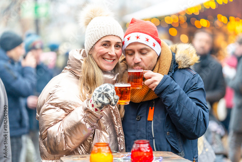 Smiling couple with mulled wine and punch mugs standing before a Christmas market glowing with holiday lights