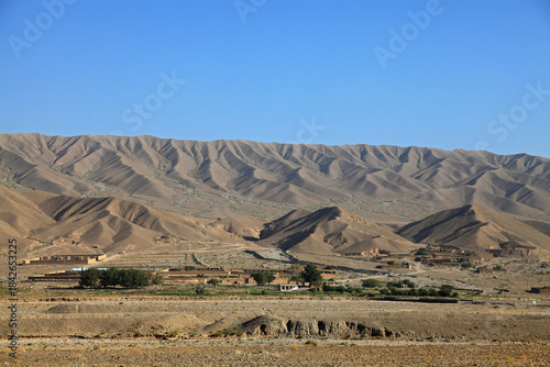 View of arid mountains rise dramatically behind a sparse village with muted buildings and scarce greenery under a clear blue sky, Quetta, Balochistan, Pakistan.
