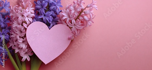 A pink heart cutout rests beside clusters of purple and pink hyacinth flowers on a pink backdrop