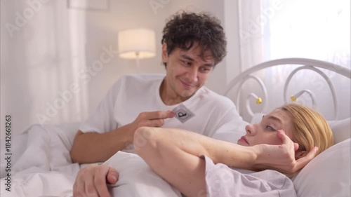 Young couple in bed holding a condom, showing intimacy, romance, and safe sex awareness in a loving relationship