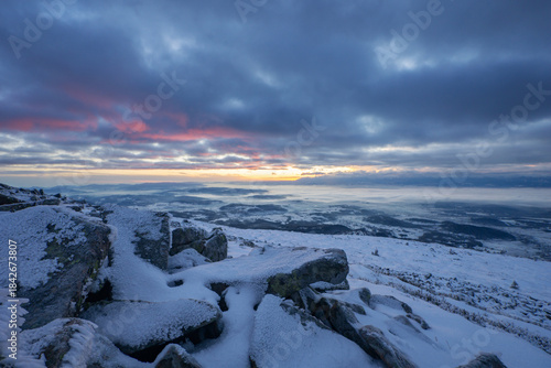 View of snow-laden rocks in the foreground contrasting with the soft pink and blue hues of the sky above the distant, misty horizon, Babia Hora, Zilina Region, Slovakia.