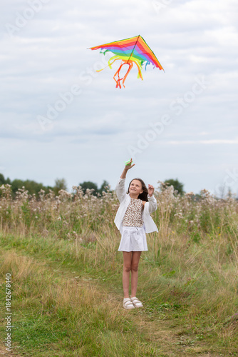 Young girl enjoys flying a colorful kite in a grassy meadow under an overcast sky during a peaceful afternoon