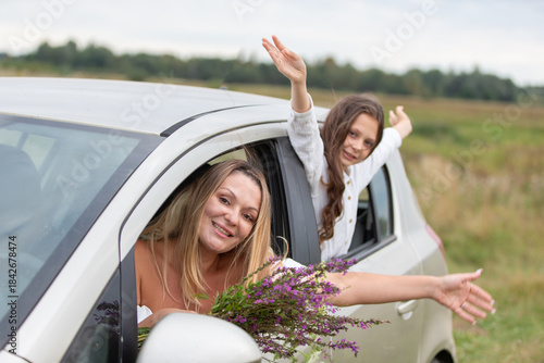 Two cheerful women enjoying a road trip while holding flowers and waving from the car on a sunny day