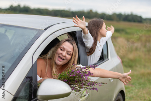 Two friends enjoy a joyful ride in a car while holding flowers and embracing the fresh air in a countryside setting