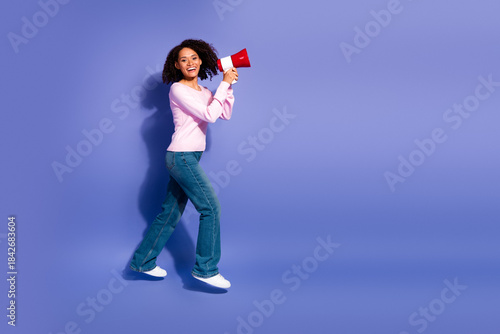Young woman with pink sweater jeans and white sneakers speaks into a red megaphone against a purple studio backdrop