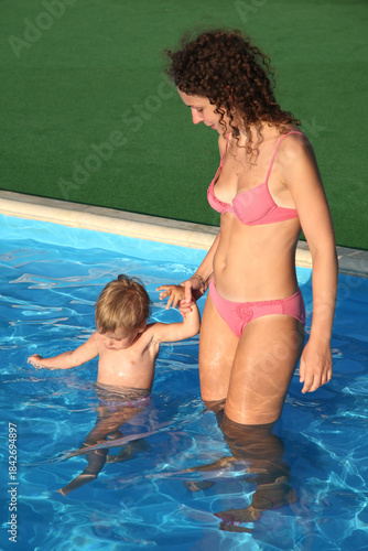 A woman is holding a child in a pool. The woman is wearing a pink bikini. The child is in the water and the woman is holding the childs hand