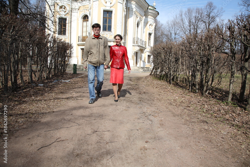 Obraz premium A man and woman are walking down a path in front of a large building. The man is wearing a jacket and jeans, while the woman is wearing a red dress. Scene is romantic and peaceful