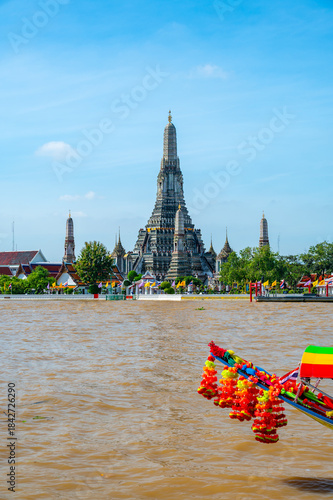 View of Wat Arun across the Chao Phraya River in Bangkok, Thailand, captured on a bright sunny day with the temple iconic spires rising above the shimmering water.