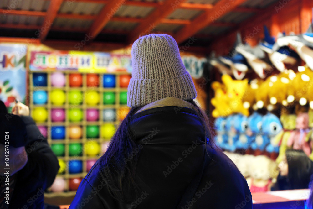 Fototapeta premium Woman wearing a knitted hat stands in front of colorful game stalls at a festive street fair during the evening, enjoying the vibrant holiday atmosphere and fun activities