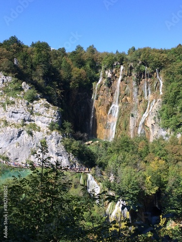 Majestic Veliki Slap Waterfall and Cascading Waterfalls in Plitvice Lakes National Park, Croatia, Surrounded by Lush Green Forest and Limestone Cliffs on a Sunny Day with Tourists on Boardwalk
