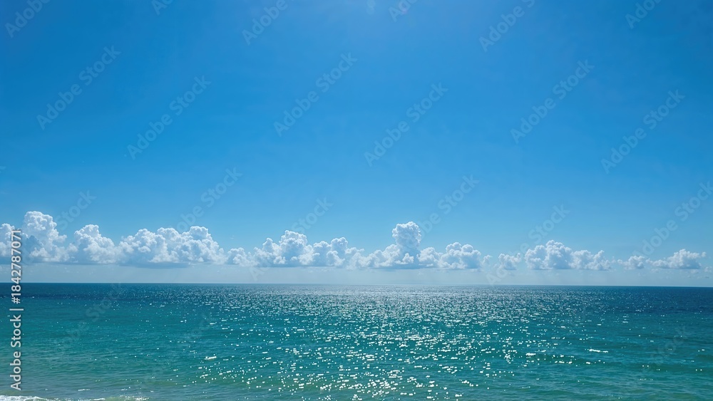Fototapeta premium A serene ocean view with a bright blue sky, scattered clouds, and sunlight reflecting off the water.
