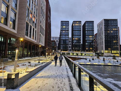 Man and a woman walking on a snowy sidewalk. The man is wearing a backpack. The sidewalk is covered in snow