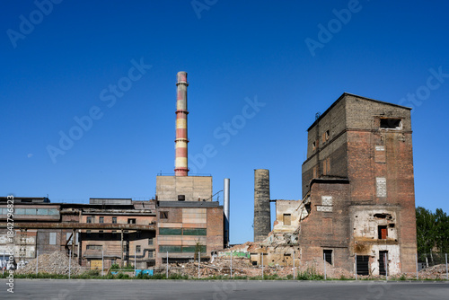 An abandoned factory with pipes, dilapidated brick buildings and pieces of walls under a clear blue sky