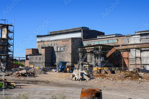 An abandoned factory with pipes, tractor, dilapidated brick buildings and pieces of walls under a clear blue sky