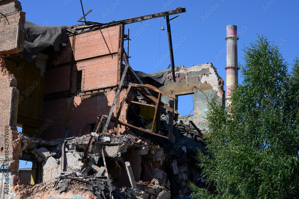 Fototapeta premium An abandoned factory with pipes, tree, dilapidated brick buildings and pieces of walls under a clear blue sky