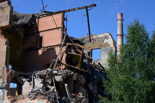 An abandoned factory with pipes, tree, dilapidated brick buildings and pieces of walls under a clear blue sky