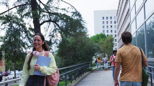 Young man and woman greeting each other with a smile while walking on the university campus, happy students enjoying a beautiful day after class with friends in the background