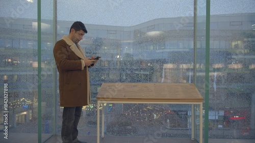 Wallpaper Mural A man in a brown shearling coat stands indoors by rain-covered windows, looking at his phone. Behind him, a softly lit shopping mall or city square is visible, with a wooden information table nearby. Torontodigital.ca