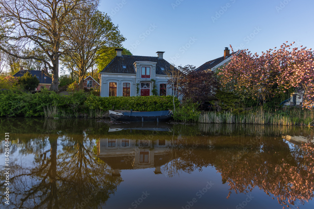 Obraz premium Traditional Dutch house by a canal in Bedum, Netherlands, with bright red windows and door. Boat, blossoming tree, and reflection in the water in warm evening light