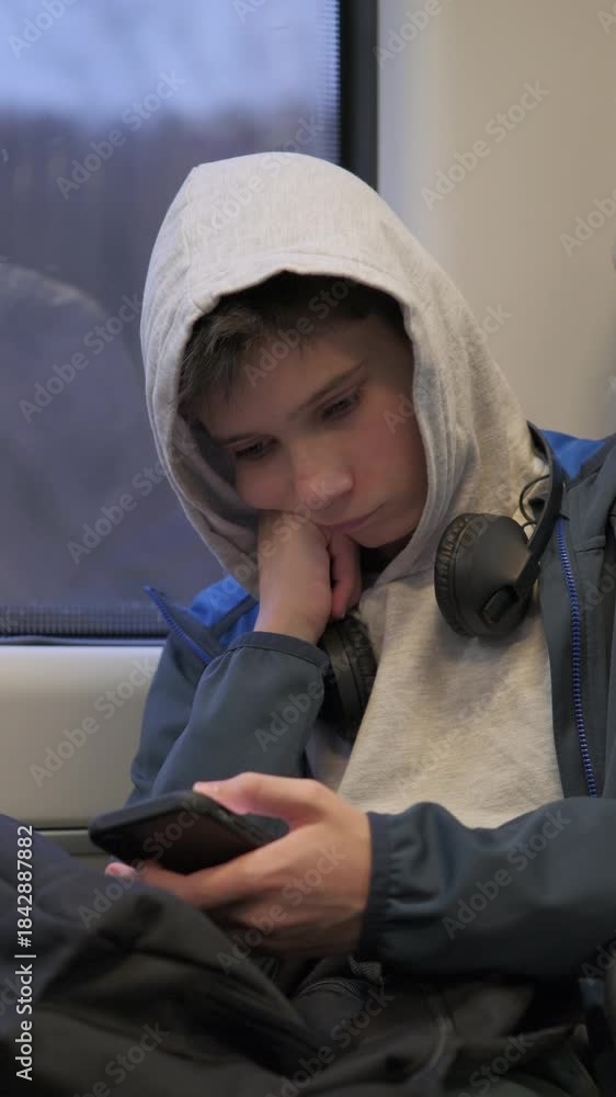 Vertical video of a bored teenage boy with hood up sitting on a train ...