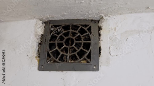 Low angle close-up shot of a dusty grey ventilation grille mounted on a rough white wall. The circular patterned vent is clogged with thick dust, cobwebs, and grime, indicating a lack of cleaning and