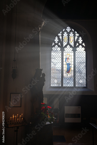 Norwich, Norfolk, UK: Church interior with a statue of Mary and Jesus. Sunlight streams through the window. Candles and flowers complete the scene.