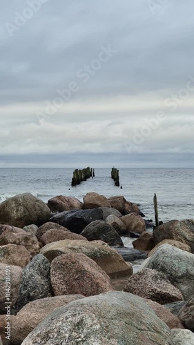 Vertical shot of rocky shoreline and remains of an old wooden pier in the calm sea. Natural ocean ambience with real sound, cloudy sky, peaceful coastal mood. 4K video.