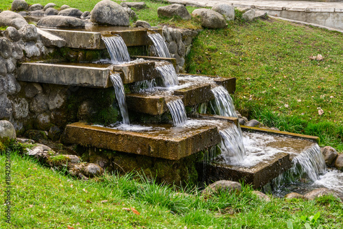 A charming, man-made tiered waterfall where clear water cascades over concrete steps, surrounded by rough stones and vibrant green mossy grass in a public garden in Razlog, Bulgaria.